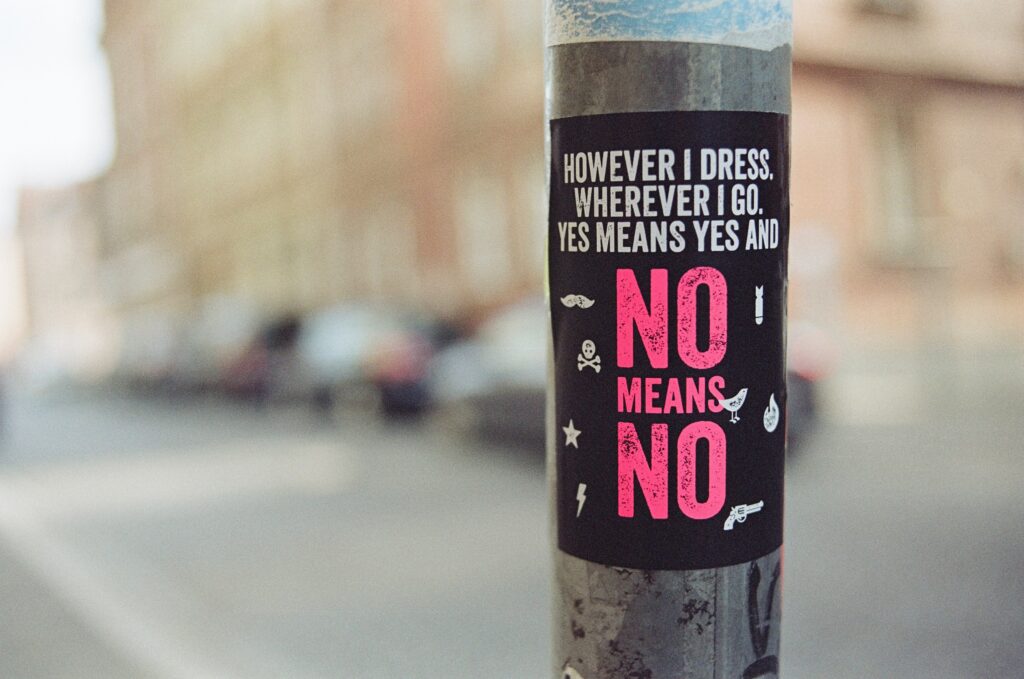 Street poll with a sign reading 'HOWEVER I DRESS, WHEREVER I GO, YES MEANS YES AND NO MEANS NO', emphasizing consent and the importance of understanding assault laws."